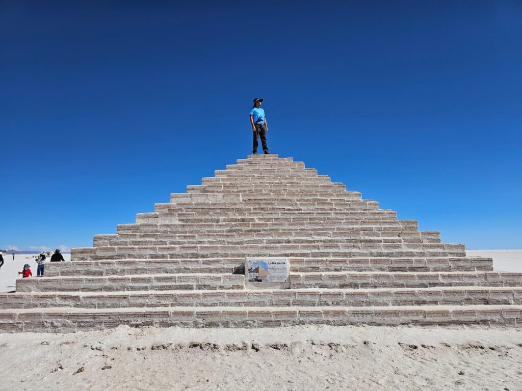 Imagem mostra turista no topo de uma pirâmide que compõe um dos belos cenários no maior deserto de sal do mundo.